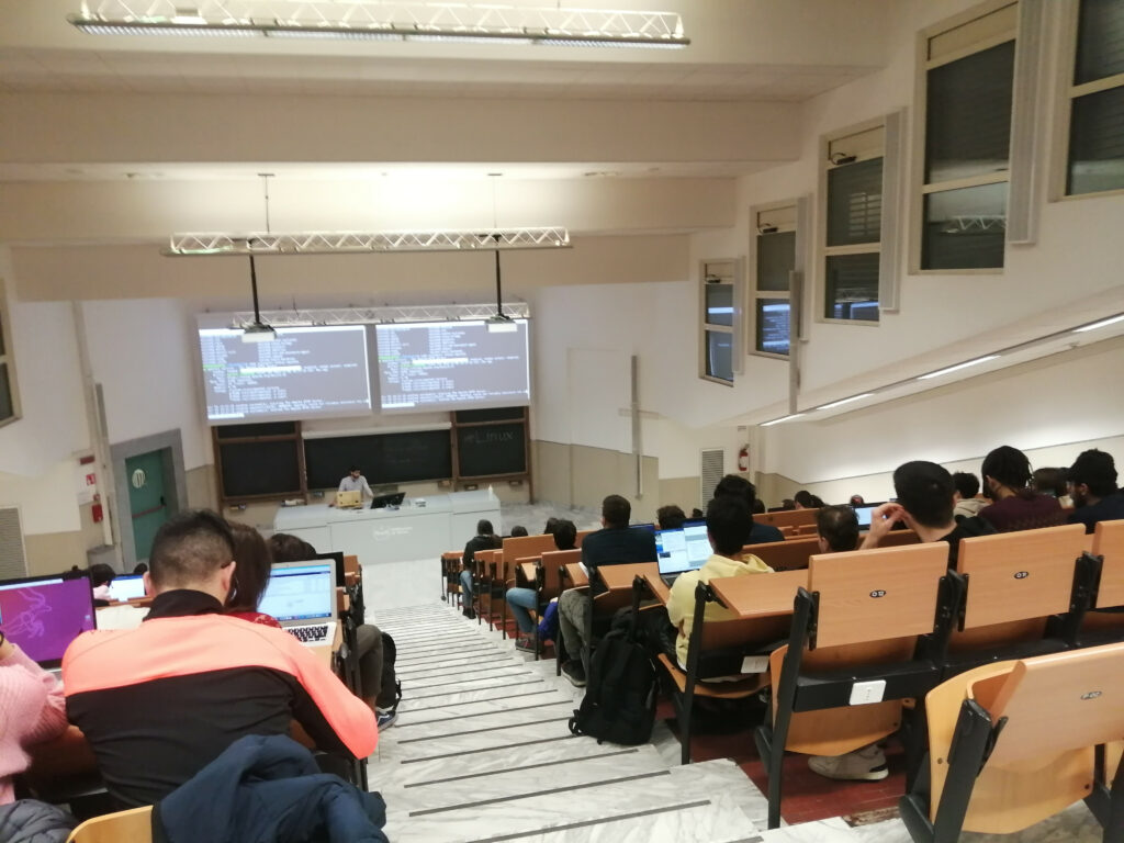 Fotografia di un'aula del Politecnico di Torino con molte persone viste di schiena mentre sui loro monitor c'è GNU/Linux. In fondo all'aula il proiettore mostra due schermi con un terminale Bash e una schermata di status di systemd. 